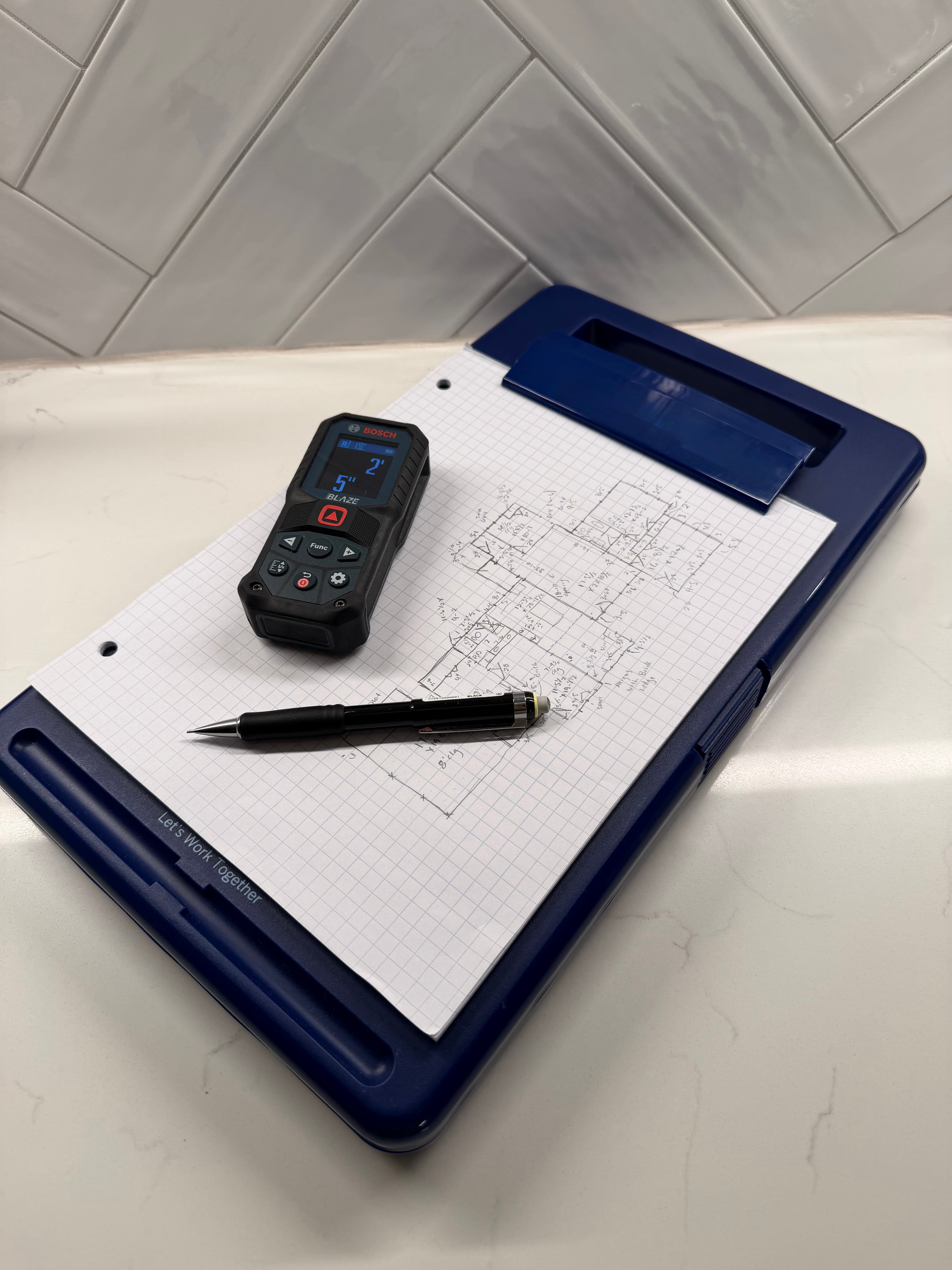 Bosch laser measurer and pencil on a blue clipboard with hand-drawn architectural floor plans.